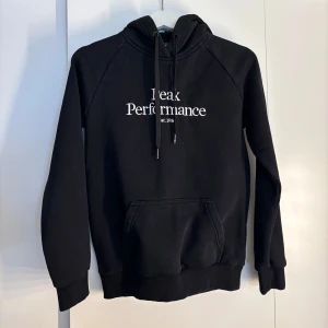 Svart hoodie från Peak Performance - Svart hoodie från Peak Performance med vit broderad logga på bröstet. Klassisk känguruficka framtill, snörning i huvan och ribbade muddar vid ärmslut och nederkant. Längst ner på snörningen är det lite skadat. 