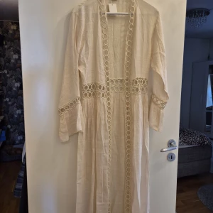 Strandklänning/kimono - Säljer en lång, ljusbeige klänning i bomull med snygga spetsdetaljer i form av cirklar längs midjan, ärmsluten och framtill. Klänningen har lång ärm och ett luftigt, bohemiskt snitt som ger en chill vibe. Perfekt för sommarens alla tillfällen! Köpt i Cypern, one size. Aldrig använd, lappar kvar.