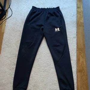 Svarta mjukisbyxor från Under Armour - Svarta mjukisbyxor från Under Armour i storlek M. Byxorna har resår i midjan, två sidofickor och en vit logga på låret. Tillverkade i 100% polyester för en sportig och bekväm känsla. Perfekta för chill eller träning.