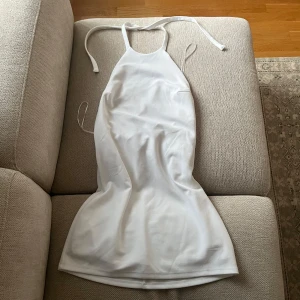 Vit halterneckklänning  - Supersnygg vit kort klänning från BikBok med halterneck och öppen rygg. Klänningen har en lätt A-linjeformad siluett och knytband i nacken. Perfekt för sommarens alla tillfällen. Materialet är mjukt och följsamt, vilket gör den både bekväm och stilren.