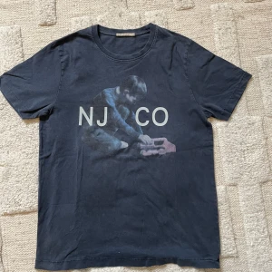 Mörkgrå t-shirt från Nudie Jeans Co - Mörkgrå t-shirt från Nudie Jeans Co med tryck av ett barn som leker med en leksak och texten 'NJ CO' på bröstet. Klassisk rund hals och korta ärmar. Skön och mjuk bomullskvalitet, perfekt för en avslappnad stil.