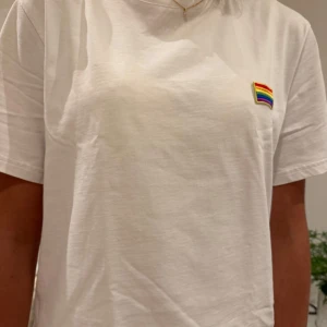 Vit t-shirt från an international anthem  - Vit t-shirt från an international anthem i storlek S. T-shirten har ett litet broderat regnbågsmärke på bröstet och klassisk rund hals. Tillverkad i mjuk bomull och har korta ärmar.