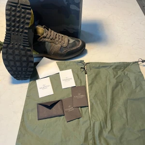 Valentino Garavani Rockrunner  - Säljer ett par Valentino Garavani Rockrunner sneakers i grått och svart med camouflage-mönster. Skorna har detaljer i mocka, skinn och textil, samt gul och vinröd häl med nitar. Klassisk snörning och platt sula. Kommer med originalpåse och box. Skick 9/10 storlek 40 men passar 41. Kvitto finns med 