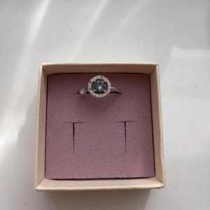 Silverring med blå sten och kristaller från Lily & Rose -  silverring med öppen design, med en rund blå sten i mitten omgiven av gnistrande stenar/kristaller( 3 av dem har lossnat) Ringen har en tunn, pärlad ringband och en extra liten kristall på sidan. Ringens storlek är justerbar och passar därför alla. 