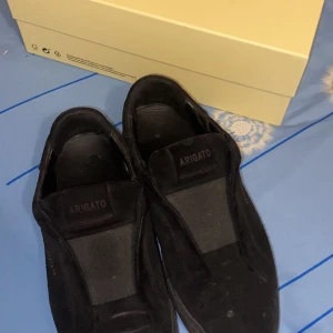 Svarta sneakers från Axel Arigato - Säljer ett par svarta sneakers från Axel Arigato. Skorna har en platt sula och är tillverkade i mocka. Perfekt för dig som gillar stilrena och moderna skor. Lite slitna men fortfarande användbara du får även med boxen och allt annat som kommer med som påsen 