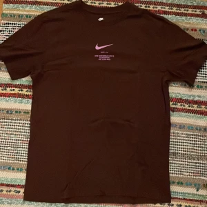 Brun Nike t-shirt med lila tryck - Snygg brun t-shirt från Nike med lila Swoosh-logga och text både fram och bak. Klassisk passform med korta ärmar och rund hals.