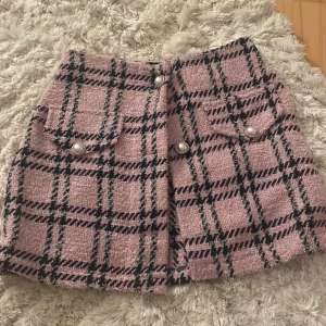 Rutig rosa kortkjol från Tezenis - Fråga om den är kvar. Superfin rosa kortkjol från Tezenis med svart och grått rutmönster. Kjolen har två fickor framtill med pärlknappar och en snygg struktur i tyget. Perfekt för dig som gillar en chic och trendig look. ( den saknas 1 knapp och den ena av de på bilden är lite sönder) 