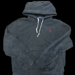 Svart hoodie från Polo Ralph Lauren - Svart hoodie från Polo Ralph Lauren med klassisk huva och vit snörning. Tröjan har en röd broderad logga på bröstet och en stor magficka. Perfekt för chill dagar och enkel att matcha med jeans eller joggers.