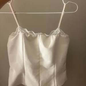 Vit korsett-topp med spetsdetalj - Säljer en stilren vit korsett-topp med tunna axelband och vacker spets längs urringningen. Toppen har en lätt strukturerad form och är perfekt för dig som vill ha en chic och feminin look. Materialet känns mjukt och har en lätt glans.