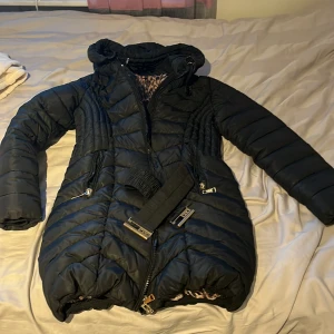 Svart lång pufferjacka Garoff med bälte - Säljer en svart lång pufferjacka från Garoff med snyggt bälte i midjan och avtagbar äkta päls till huvan. Jackan har dragkedjor, två fickor med päls inuti och ett coolt leopardmönstrat innerfoder. Perfekt för kalla dagar när du vill vara både varm och stylish. Storlek 38. Om man vill köpa UTAN PÄLS då sänks priset. Men ni måste kontakta mig för det. (Skriv för detaljerade bilder). 