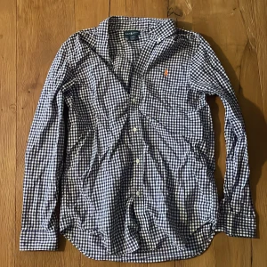 Ralph Lauren skjorta - Snygg rutig skjorta från Ralph Lauren i blått och vitt med klassisk krage och knäppning framtill. Skjortan har lång ärm och den ikoniska orangea Polo-loggan broderad på bröstet. Perfekt för dig som gillar stilrena och tidlösa plagg. Storlek 18-20 teen, så sitter som en vanlig S.