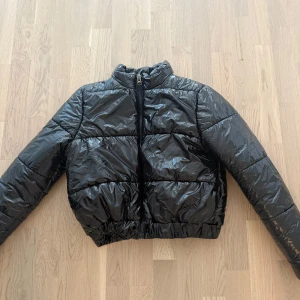 Svart pufferjacka från Chiquelle XS - Säljer en svart, glansig pufferjacka från Chiquelle i storlek XS. Jackan har en croppad passform, hög krage och dragkedja framtill. Perfekt för dig som vill ha en trendig och varm jacka med snygg streetkänsla.