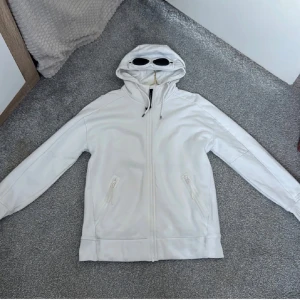 Vit Goggle-hoodie från C.P. Company - Vit hoodiejacka från C.P. Company med ikoniska svarta goggle-linser i huvan. Jackan har dragkedja framtill, två fickor med dragkedja och justerbar huva med snörning. Tillverkad i mjuk bomull för en clean och modern look.