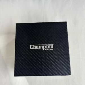 Champion Spider Sport herrklocka brun - Snygg herrklocka från Champion Spider Sport med mörkgrå urtavla, roséguldsdetaljer och datumvisning. Klockan har ett klassiskt runt silverfärgat boett och ett brunt läderarmband. Levereras i originalbox.