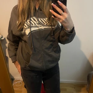 Mörkgrå hoodie med tryck och dragkedja - Säljer en mörkgrå hoodie med vitt tryck över bröstet. Det står NewYork. Tröjan har huva med snörning, dragkedja framtill och stora fickor. 