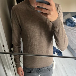Merinoull tröja från Hugo boss  - Slim fit tröja från Hugo Boss i extrafin merinoull. Snygg beige färg, rund halsringning och diskret metallogo på sidan. Perfekt för dig som gillar stilrena och bekväma plagg med en exklusiv känsla.