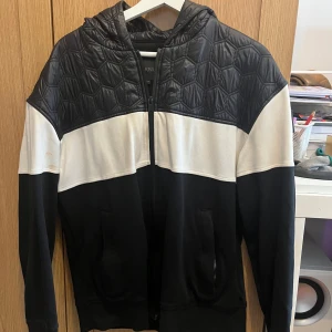 Svartvit hoodie från Moose Knuckles - Säljer en snygg hoodie från Moose Knuckles med svart quiltad huva och axelparti, vit mittsektion och svarta ärmar och nederdel. Tröjan har dragkedja framtill, två fickor och en tydlig logga på ärmen. Perfekt för dig som gillar streetwear med coola detaljer.