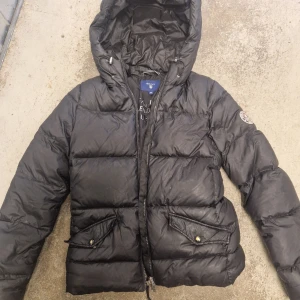 Svart pufferjacka från Gant XS - Säljer en svart pufferjacka från Gant i storlek XS. Jackan har huva med snörning, dragkedja framtill och två stora fickor med lock och knapp. Insidan har en innerficka med dragkedja och snygga detaljer längs fodret. 