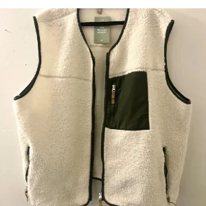 Teddyväst  - Mysig beige fleeceväst från H&M med svarta detaljer och dragkedja framtill. Västen har en cool grön bröstficka med dragkedja och två sidofickor. Perfekt att slänga över en hoodie eller stickad tröja för extra värme.