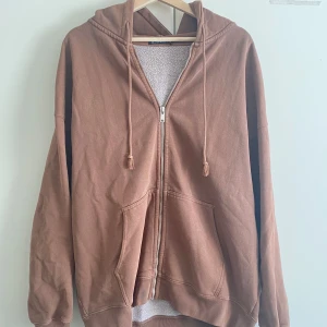Brun hoodie med dragkedja Brandy Melville - Mysig brun hoodie från Brandy Melville med dragkedja framtill, huva och snörning. Tröjan har två stora fickor och är i en loose passform. Perfekt för chill dagar och passar till allt. Tillverkad i mjuk bomullsmix. Har någon fläck som förmodligen går bort i tvätten