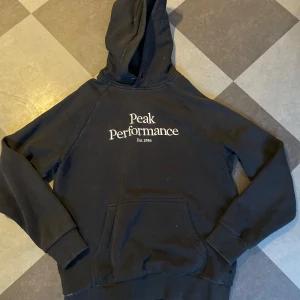 Svart hoodie från Peak Performance - Svart hoodie från Peak Performance med vit broderad logga på bröstet. Känguruficka framtill och huva. Använd och några få tecken på användning längst ned vid tröjmudden