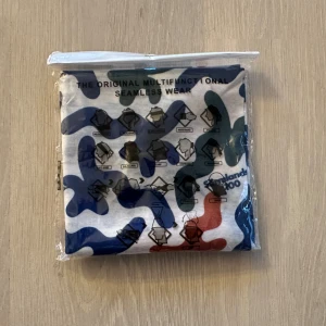 Multifunktionell huvudbonad Sörmlands 100 - Säljer en multifunktionell huvudbonad från Sörmlands 100 med coolt camouflage-mönster i blått, grönt och rött på vit botten. 