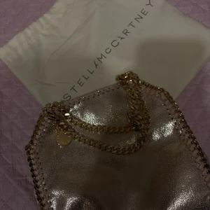 Glittrig axelväska Stella McCartney - Snygg glittrig axelväska från Stella McCartney i ljusrosa med kedjedetaljer i guld. Väskan har en cool metallisk finish och en rund logotag på kedjan. Perfekt för dig som vill sticka ut med en trendig accessoar.