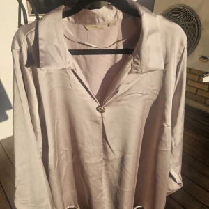 Champagnefärgad satinblus från MyMuse - Snygg champagnefärgad blus i satin från MyMuse med klassisk krage och en dekorativ knapp framtill. Blusen har lång ärm och en lyxig, glansig finish som ger en elegant vibe. Perfekt för dig som gillar stilrena och trendiga plagg.