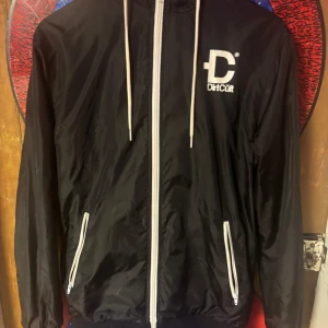 Svart vindjacka från DirtCult - Svart vindjacka från DirtCult med vit dragkedja och vita detaljer. Jackan har huva med snörning, två fickor med dragkedja och meshfoder på insidan. Fläckig på insidan å i luvan, även ett hål på toppen av luvan. Fläckarna går säkert bort i tvötten. 