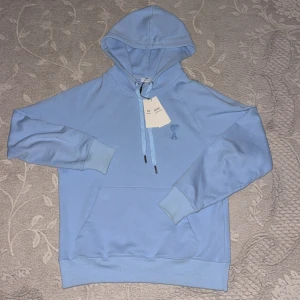 Ljusblå hoodie från Ami Paris - Säljer en stilren ljusblå hoodie från Ami Paris med broderad logga på bröstet och snörning i huvan. Tröjan har känguruficka och ribbade muddar. Perfekt för dig som gillar en clean och avslappnad look.