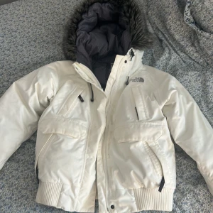 Vit pufferjacka från The North Face - Säljer en snygg vit pufferjacka från The North Face med en stor huva och fuskpäls. Jackan har dragkedja, stora fickor framtill och ribbade muddar. Perfekt för kalla dagar och riktigt skön att bära. Klassisk logga på bröstet och cool passform.