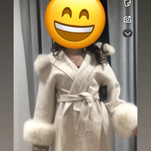 Beige kappa - Säljer en superfin beige kappa med fluffiga pälsdetalljer på huva och ärmslut. Kappan har knytbälte i midjan, stora fickor framtill och en snygg omlottmodell. Perfekt för dig som vill ha en mysig och trendig look under höst och vinter! Använd endast 1 säsong och köptes för 3500. En av armarna hade tappat en knapp som jag sytt tillbaka.