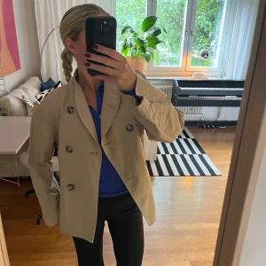 Beige jacka med dubbelknäppning - Säljer en beige kavaj med dubbelknäppning och stora, mönstrade knappar. Jackan har en rak och lite oversized passform med långa ärmar och klassisk krage. Tun jacka som passar nu i början av hösten! Säljer jackan till MYCKET nedsatt pris då den har en fläck i ryggen. Inte särskilt märkbar vidbedsläppt hår (sitter högt upp, se bild) 