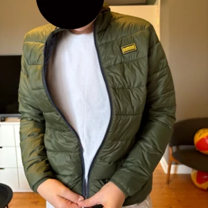 Olivgrön pufferjacka från Barbour - Snygg olivgrön pufferjacka från Barbour International med quiltad design och svart dragkedja. Jackan har en klassisk passform, hög krage och Barbour-logga på bröstet. Perfekt för kyliga dagar och enkel att matcha med olika outfits.