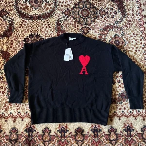 Svart stickad tröja från Ami Paris - Svart stickad tröja från Ami Paris med rund hals och ribbade muddar. På bröstet finns ett stort rött hjärta med bokstaven A, vilket ger en unik och trendig look. Perfekt för dig som vill sticka ut med en ikonisk detalj.