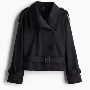 trenchcoat  - Stilren svart kort trenchcoat med dubbelknäppning, bred krage och bälte i midjan. Jackan har långa ärmar med spännen vid ärmslut och axelklaffar för en klassisk look. Perfekt för dig som gillar en tidlös och cool stil.