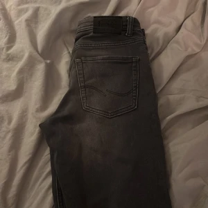 Svarta jeans från Jack & Jones - Säljer ett par svarta jeans från Jack & Jones med klassisk femficksdesign och svarta sömmar. Jeansen har en rak passform och är tillverkade i ett mjukt bomullsmaterial med lite stretch för extra komfort. Perfekta för dig som gillar en stilren look. Litet hål som är igensytt men annars jättebra skick. Tveka inte på att höra av dig vid frågor!
