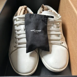 Vita Saint Laurent sneakers - Snygga vita sneakers från Saint Laurent Paris i stilren design. Skorna är tillverkade i slätt skinn med rund tå, platt sula och klassisk snörning. Perfekta för dig som gillar minimalistisk och lyxig streetstyle. Kommer med originalbox och extra snöre. Skorna har använts fåtal gånger och är i bra skick, strolek 39 men pasar mig som har 40,5!