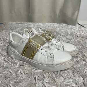 Valentino Rockstud vita sneakers - Snygga vita Valentino Rockstud sneakers med guldiga detaljer och nitar. Skorna har en bred guldig rem över vristen med ikoniska pyramidformade nitar och klassisk snörning. Tillverkade i skinn med platt sula och rund tå. Perfekta för dig som vill sticka ut med en lyxig touch.