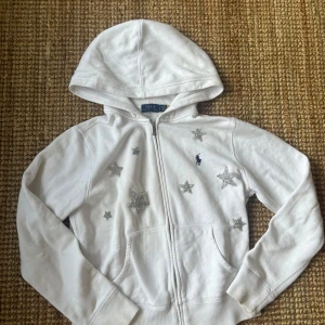 Vit hoodie från Polo Ralph Lauren med stjärnor - Vit hoodie från Polo Ralph Lauren med glittriga silverstjärnor på framsidan. Tröjan har dragkedja, huva och två fickor framtill. Klassisk blå logga broderad på bröstet. Perfekt för dig som vill sticka ut med lite extra bling. Kolla gärna min profil och skriv ifall frågor uppkommer eller om du vill se fler bilder ❣️❣️