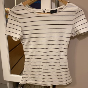 Randig topp från Vila i bomull XS - Vit topp från Vila med svarta horisontella ränder. Modellen är figurnära med korta ärmar och rund halsringning. Dragkedja bak för en snygg detalj. Perfekt till jeans eller kjol för en enkel och stilren look.