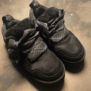 Uggs - Säljer ett par svarta sneakers från UGG med fluffig päls på insidan och grova snören. Skorna har en sportig look med mesh och mockadetaljer, samt en rejäl platt sula. Perfekta för dig som vill ha både stil och komfort under kyligare dagar. Nypris gick för 3 tusen kronor
