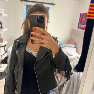 Svart skinnjacka med fransar - Säljer en svart skinnjacka med coola fransdetaljer längs ärmar och rygg. Jackan har klassisk bikerstil med dragkedja framtill och markerade axlar. Perfekt för dig som vill sticka ut och addera lite edge till din outfit.