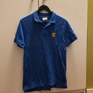 Blå pikétröja med Porsche-logga - Snygg blå pikétröja med klassisk krage och Porsche-emblem på bröstet. Kortärmad modell i mjuk bomull, perfekt för dig som gillar sportig och stilren look. Passar till jeans eller shorts för en avslappnad vibe.