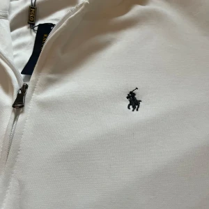 Vit hoodie från Polo Ralph Lauren - Vit hoodie från Polo Ralph Lauren i mjuk bomull. Klassisk design med huva, dragkedja och två fickor fram. Ikonisk Polo-logga broderad på bröstet i svart. Perfekt för chill dagar och enkel att matcha med jeans eller joggers. Använd 1 gång, var förliten för mig.