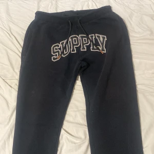 Svarta mjukisbyxor Supply & Demand L - Svarta mjukisbyxor från Supply & Demand med broderad SUPPLY-text framtill och snygga detaljer på snörningen. Byxorna har elastisk midja med dragsko och tryckdetalj på benet. Perfekta för en chill och avslappnad stil.