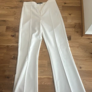 Vita kostymbyxor från Gina Tricot S - Snygga vita kostymbyxor från Gina Tricot i storlek S. Byxorna har hög midja, raka ben med pressveck och diskret bakficka. Stängs med dragkedja och hake framtill. Perfekta för en clean och stilren look.