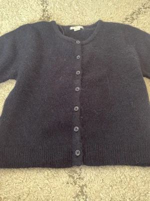 Mörkblå kofta från Gina Tricot - Säljer en mörkblå, mjuk och fluffig kofta från Gina Tricot i storlek 146/152. Koftan har rund hals, långa ärmar och knäppning med svarta knappar hela vägen framtill. Perfekt att slänga på sig när det är lite kyligt ute.