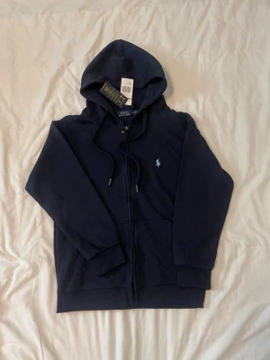 Polo Ralph Lauren zip-hoodie mörkblå – ljusblå logga (S)  - Riktigt fet mörkblå zip-hoodie från Polo Ralph Lauren med ljusblå logga. Clean look som funkar till allt jeans, cargos eller mjukis. Storlek S men passar även XS, sitter snyggt men är lite kort i armarna. Väldigt bra skick, inga hål eller fläckar. Perfekt om du vill ha en stilren men ändå snygg tröja 