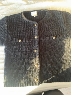 Svart stickad kofta från H&M - Kortärmad svart stickad kofta från H&M i storlek S. Koftan har rund hals, dekorativa guldknappar framtill och på fickorna samt ribbade kanter. Perfekt för lager på lager och ger en chic touch till din outfit.